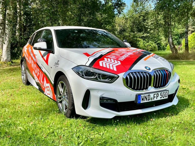 Ein weißer BMW mit Fahrschulfolierung steht auf einer Wiese.