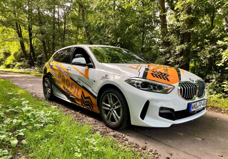 Ein weißer BMW mit Fahrschulfolierung steht auf einem Feldweg.